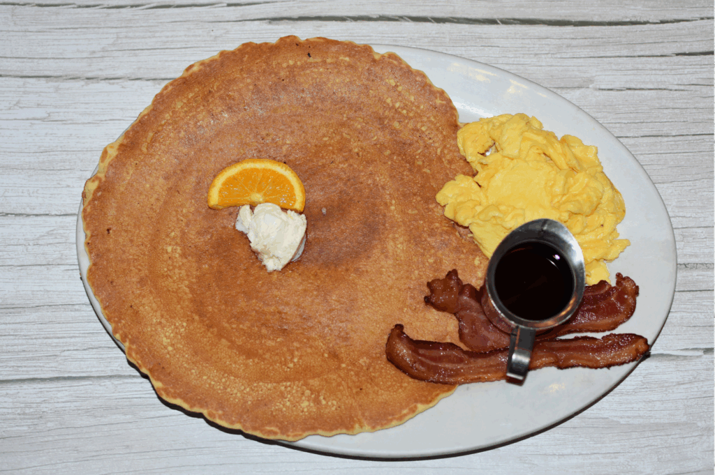 Oversized Hash House A Go Go pancake with scrambled eggs, bacon, butter, syrup, and an orange slice at Flamingo Crossings.